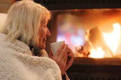 An older woman wrapped in a blanket, holding a hot drink and sitting in front of a fire.