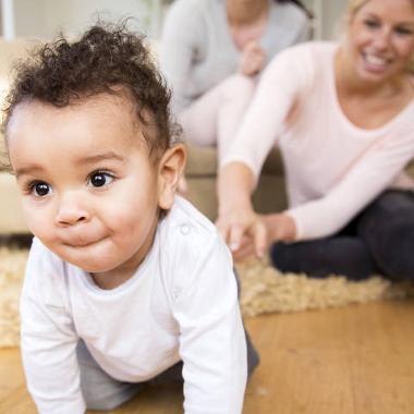 Photograph of baby crawling towards the camera. Adult smiles and reaches for the child.