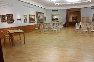 Photograph of front view of Colman Ceremony Room including wooden floor, chairs in rows, paintings in gold frames
