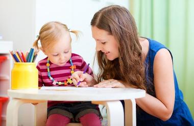 Photo of adult crouching down to help a child, who sits at a small table, draw a picture. 