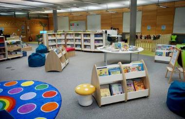 Book shelves and tables displaying children’s books, bean bags, stools and a large colourful mat to sit on