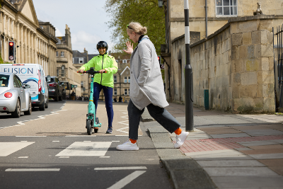 Photograph of person riding an e-scooter waiting at a zebra crossing for a pedestrian to pass