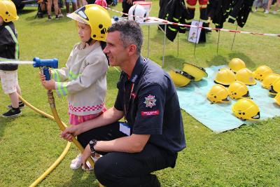 Firefighter crouching next to young girl who is wearing a helmet and using a hose