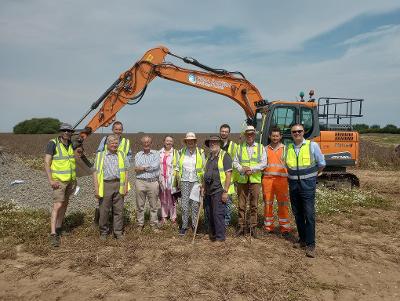 Photo of the construction site and eleven people are pictured in front of a digger. Pictured left to right: Mark Ogden – Flood & Water Manager, NCC , Hull  – Assistant Director – Waste and Water Management, NCC, Tim Goddard – Local Farmer, Sir David Ralli