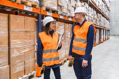 A man and a woman in a warehouse wearing hi-vis clothing and hard hats